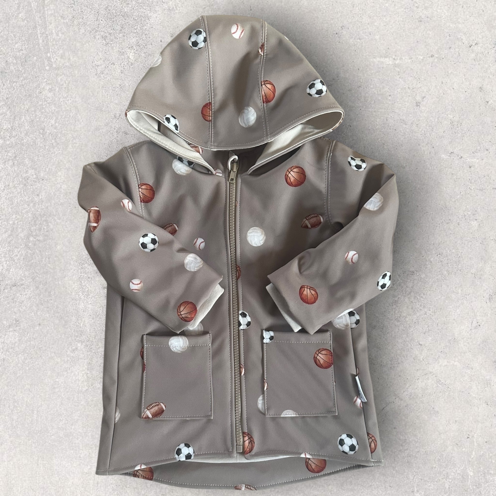 Beigegraue Kinder Softshelljacke mit  Fußbällen, Basketball- und Baseball-Motiven, Kapuze und hohem Kragen.
